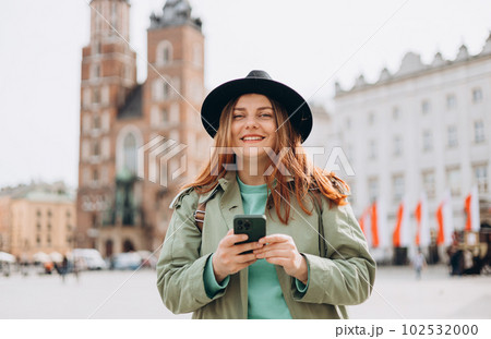 Beautiful stylish woman in hat walking on Market Square in Krakow on autumn day and holding mobile phone. Urban lifestyle concept. Check social networks, send sms or book hotel Beautiful stylish woman in hat walking on Market Square in Krakow on autumn day and holding mobile phone. Urban lifestyle concept. Check social networks, send sms or book hotel 102532000