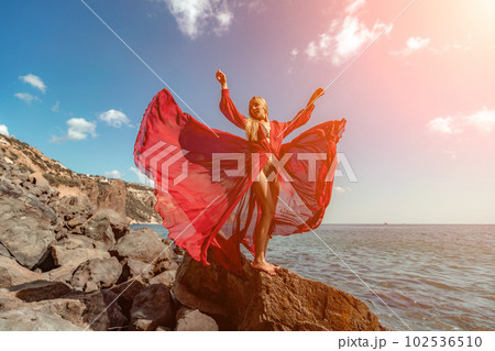 Red dress sea woman. A blonde with flowing hair in a long flowing red dress stands on a rock near the sea. Travel concept, photo session at sea Red dress sea woman. A blonde with flowing hair in a long flowing red dress stands on a rock near the sea. Travel concept, photo session at sea 102536510