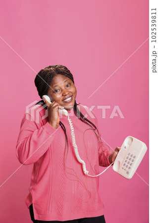 Woman with pink sweather talking on landline phone call discussing with remote friend, conversation with retro telephone cord line in studio. Model having fun speaking on vintage phone over background 102539811