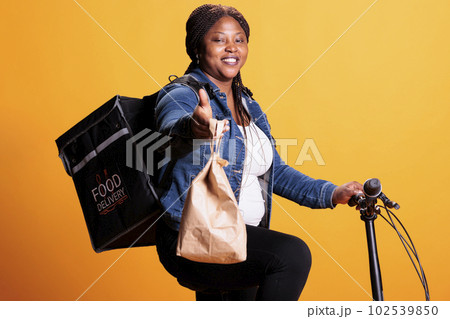 Takeaway delivery employee holding recycled paper bag delivering fast food order to customer using bike as transportation. Restaurant courier standing in studio with yellow background Takeaway delivery employee holding recycled paper bag delivering fast food order to customer using bike as transportation. Restaurant courier standing in studio with yellow background 102539850