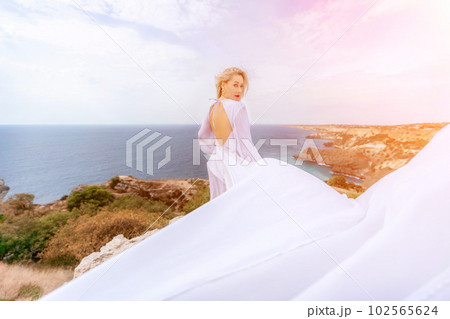 Blonde with long hair on a sunny seashore in a white flowing dress, rear view, silk fabric waving in the wind. Against the backdrop of the blue sky and mountains on the seashore. 102565624