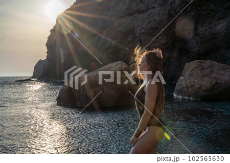 Woman tourist enjoying the sunset over the sea mountain landscape. Sits outdoors on a rock above the sea. She is wearing jeans and a blue hoodie. Healthy lifestyle, harmony and meditation 102565630