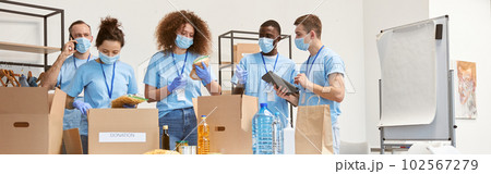 Full length shot of volunteers in protective masks and gloves sorting, packing food and water in cardboard boxes, working together on donation project indoors 102567279