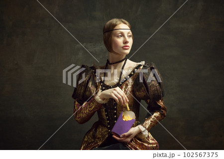 Portrait of young beautiful woman in vintage costume, eating fries against dark green background. Fast food lover 102567375