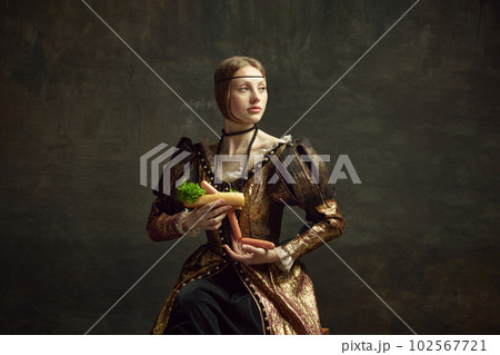 Portrait of young beautiful woman in vintage, elegant dress holding giant hot dog against dark green background. Fast food 102567721