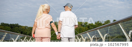 Positive senior woman and man walk holding hands along footbridge after training 102567995