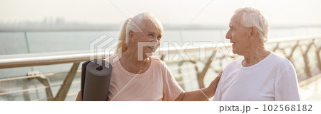 Smiling senior woman with mat cheers up man with poles for Nordic walking on footbridge Smiling senior woman with mat cheers up man with poles for Nordic walking on footbridge 102568182