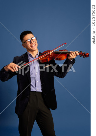 Portrait with handsome, young musicians, violinist wearing elegant suit and eyeglasses playing on violin and smiling over blue color studio background Portrait with handsome, young musicians, violinist wearing elegant suit and eyeglasses playing on violin and smiling over blue color studio background 102575023