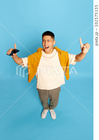 Portrait with excited young man, guy, student in stylish casual clothes listening to music via earpods and smiling over blue background. Top view Portrait with excited young man, guy, student in stylish casual clothes listening to music via earpods and smiling over blue background. Top view 102575135