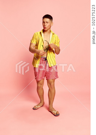 Portrait of attractive young guy, student, man wearing beach clothes and sunglasses grimacing over pink color studio background Portrait of attractive young guy, student, man wearing beach clothes and sunglasses grimacing over pink color studio background 102575223