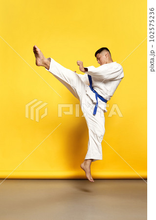 Portrait of professional male taekwondo, karate athlete wearing white kimono doing basic movements over yellow background 102575236
