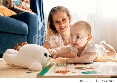 Caring joyful mother plays with her little child, lying on the floor in a bright children's room. Various toys, a book. Learning with play. Caring joyful mother plays with her little child, lying on the floor in a bright children's room. Various toys, a book. Learning with play. 102576967