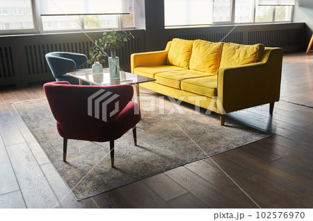 Part of the interior with a yellow sofa and 2 armchairs, blue and red, magazine table and carpet. Window on the background, decor on the table, vases and copy space. High quality photo 102576970