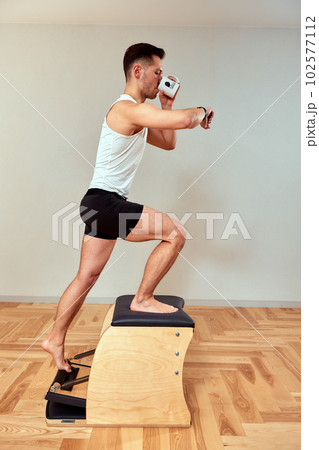 Young man during pilates fitness gymnastics course on a stepper looks at his watch and drinks coffee from a cup, pilates classes on a reformer, endurance training Young man during pilates fitness gymnastics course on a stepper looks at his watch and drinks coffee from a cup, pilates classes on a reformer, endurance training 102577112