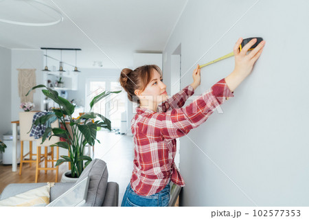Young woman doing measuring with a measure tape on the wall. Girl wants to put a picture on the wall at home. Housekeeping work. Doing repair herself. DIY, gender equality in work concept. Young woman doing measuring with a measure tape on the wall. Girl wants to put a picture on the wall at home. Housekeeping work. Doing repair herself. DIY, gender equality in work concept. 102577353