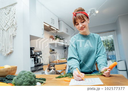 Young woman filling veganuary calendar while eating fresh carrot sitting on kitchen. Fresh vegetables and fruits on table. Vegan and vegetarian diet eating and cooking at home. Selective focus 102577377