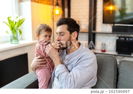 Father with baby, man sitting on sofa with toddler child playing and kissing kid's hand. Father with baby, man sitting on sofa with toddler child playing and kissing kid's hand. 102580897