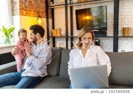 Father carrying infant daughter mother using laptop remote work husband helping with baby child Father carrying infant daughter mother using laptop remote work husband helping with baby child 102580898