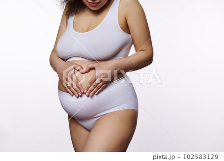 Close-up of a pregnant woman in white lingerie, expecting baby, enjoying her 30 week of pregnancy, making a heart shape with hands on her belly, isolated white background. Femininity Maternity Nudity 102583129