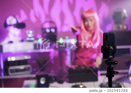 Asian dj with pink hair performing techno music using professional mixer console while recording process with mobile phone camera. Smiling artist playing song during night time in club 102591288