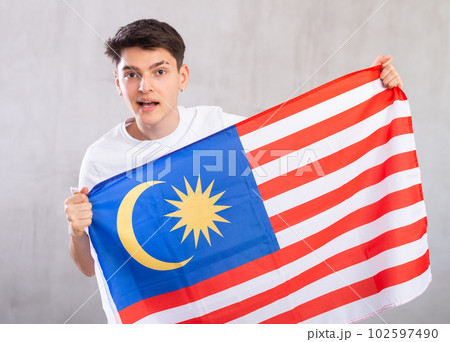 Happy young man holding flag of Malaysia against unicoloured background Happy young man holding flag of Malaysia against unicoloured background 102597490