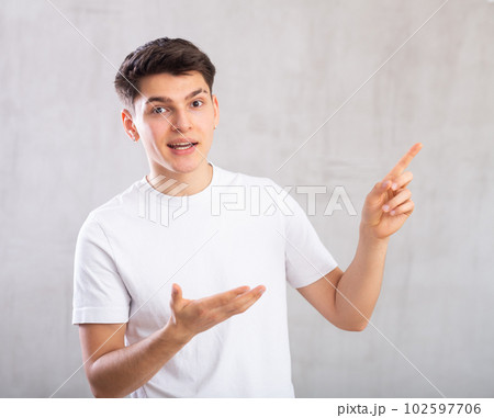 Surprised young man with pointing hand gestures standing against background of light unicolored wall 102597706