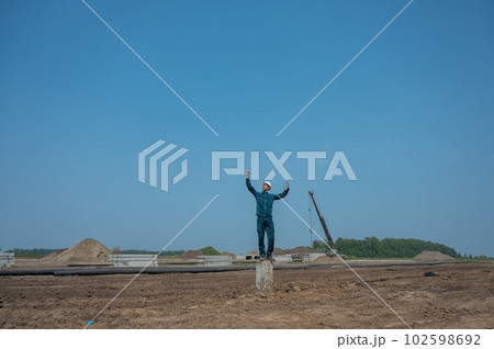 Builder in a protective helmet holds a laptop and stands on a pile in the middle of a construction site. Builder in a protective helmet holds a laptop and stands on a pile in the middle of a construction site. 102598692