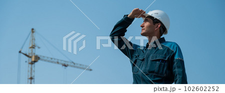 A builder in work clothes and a helmet stands on a construction site against the background of a construction crane. A builder in work clothes and a helmet stands on a construction site against the background of a construction crane. 102602252