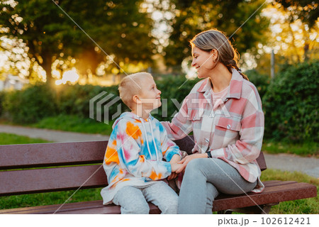 mother and son sit on a park bench in the rays of the setting sun. the concept of a family. Mother's Day. beautiful girl (mother) with a boy (son) in the park in the park are sitting on a bench at 102612421