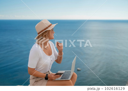 Freelance women sea working on the computer. Good looking middle aged woman typing on a laptop keyboard outdoors with a beautiful sea view. The concept of remote work. 102616129