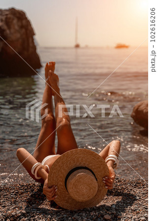 woman sea travel Tanned middle-aged woman with long hair and a white bathing suit. He sits on the seashore in a large sun hat with his back and looks at the sea. woman sea travel Tanned middle-aged woman with long hair and a white bathing suit. He sits on the seashore in a large sun hat with his back and looks at the sea. 102616206