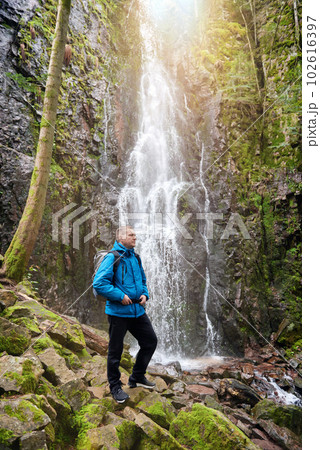 Tourist attraction of Germany - falls of Burgbach Waterfall near Schapbach, Black Forest, Baden-Wurttemberg, Germany. Man hiker in blue jacket standing on stone and looks at flow of falling water Tourist attraction of Germany - falls of Burgbach Waterfall near Schapbach, Black Forest, Baden-Wurttemberg, Germany. Man hiker in blue jacket standing on stone and looks at flow of falling water 102616397