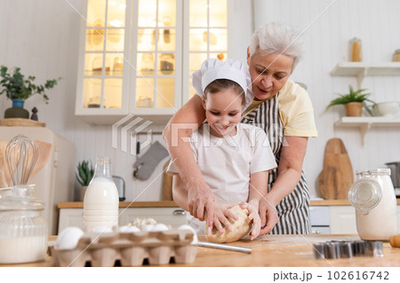 Happy family in kitchen. Grandmother and granddaughter child cook in kitchen together. Grandma teaching kid girl knead dough bake cookies. Household teamwork helping family generations concept Happy family in kitchen. Grandmother and granddaughter child cook in kitchen together. Grandma teaching kid girl knead dough bake cookies. Household teamwork helping family generations concept 102616742