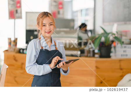 Portrait of confident female barista standing at counter. Woman cafe owner in apron looking at camera and smiling while welcoming guests. Portrait of confident female barista standing at counter. Woman cafe owner in apron looking at camera and smiling while welcoming guests. 102619437
