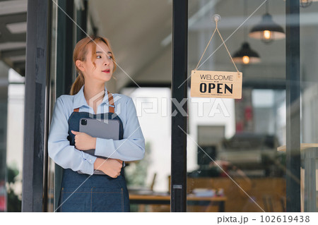 Portrait of confident female barista standing at counter. Woman cafe owner in apron looking at camera and smiling while welcoming guests. Portrait of confident female barista standing at counter. Woman cafe owner in apron looking at camera and smiling while welcoming guests. 102619438