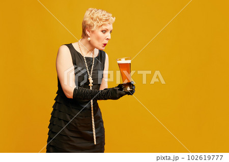 One surprised blond mature woman in vintage black dress holding beer glass and looking on it over yellow background. Treasure 102619777