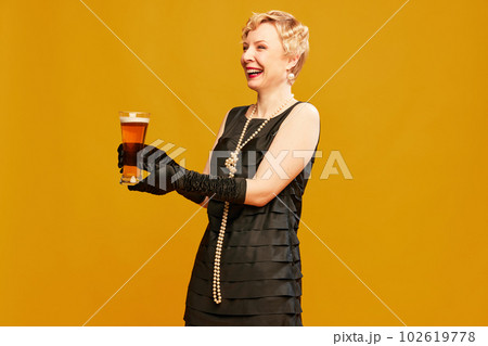 Portrait of happy blond mature woman in retro black dress holding beer glass and smiling over yellow studio background. Alcohol degustation Portrait of happy blond mature woman in retro black dress holding beer glass and smiling over yellow studio background. Alcohol degustation 102619778