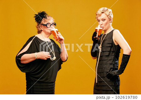 Portrait with two women senior and mature in vintage clothes drinking foamy lager beer over yellow background. Fresh beer draft 102619780