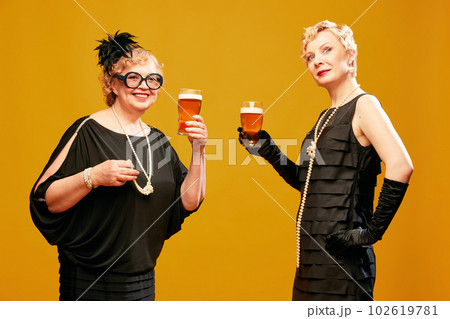 Portrait with attractive two women, friends wearing black dresses enjoying fresh beer over yellow background. Female party 102619781