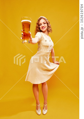 Smiling pretty girl, waitress in retro dress holding beer glass and serving over yellow background. Oktoberfest bavarian culture 102619784