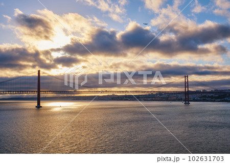 View of 25 de Abril Bridge famous tourist landmark of Lisbon connecting Lisboa and Almada on Setubal Peninsula over Tagus river on sunset with flying plane. Lisbon, Portugal. Camera pan 102631703