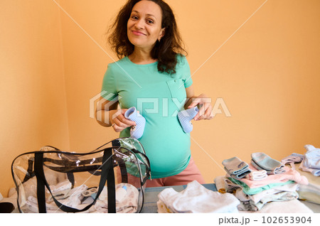 Happy smiling pregnant woman, expectant mother feeling positive emotions, expecting her newborn child, holding knitted baby boots on her belly while collecting things for the maternity hospital bag 102653904