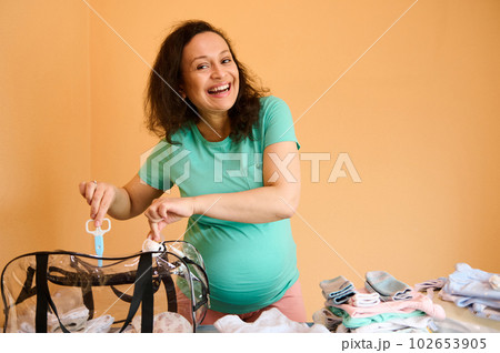 Pregnant woman smiles at camera, puts a bottle with baby body lotion and nail scissors in the bag, standing by ironing board, collecting newborn things in bag for the hospital, happy by expecting baby 102653905