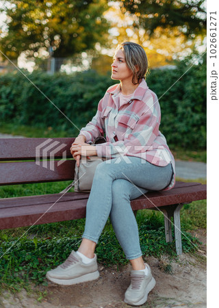 Girl enjoying city view from a bench in sunset or sunrise time. Beautiful blond-haired pretty girl in summer green park against sunset beams. Lonely woman sitting alone moments sunset. On Blurred Girl enjoying city view from a bench in sunset or sunrise time. Beautiful blond-haired pretty girl in summer green park against sunset beams. Lonely woman sitting alone moments sunset. On Blurred 102661271