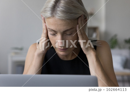 Tired mature blonde woman in casual touching head at laptop 102664113