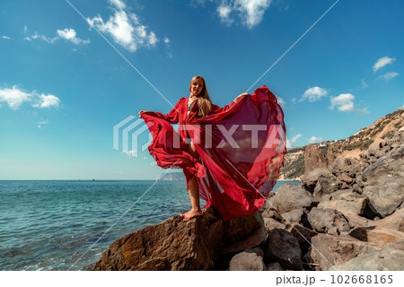 Red dress sea woman. A blonde with flowing hair in a long flowing red dress stands on a rock near the sea. Travel concept, photo session at sea Red dress sea woman. A blonde with flowing hair in a long flowing red dress stands on a rock near the sea. Travel concept, photo session at sea 102668165