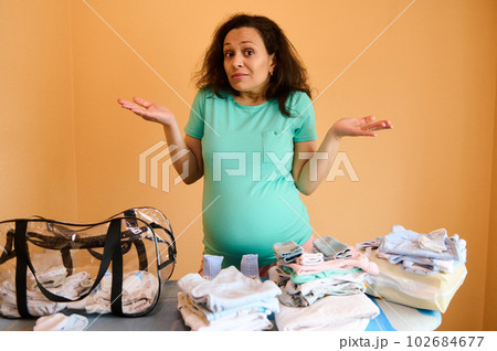 Surprised multi ethnic adult pregnant woman, standing by ironing board with clean ironed baby laundry, holding hands palms up, looking at camera, asking what to pack into a bag for maternity hospital 102684677