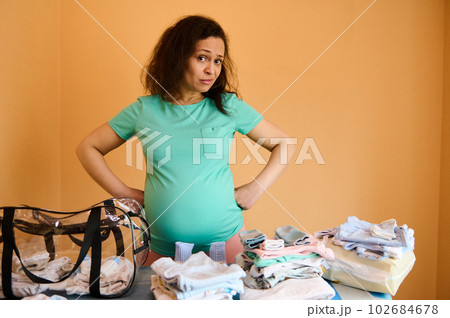 Doubtful ethnic pregnant woman packing bag for maternity hospital, standing by ironing board with a pile of clean and ironed newborn laundry. Pregnancy, maternity leave, childbirth and people concept 102684678