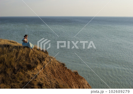 A girl in a long dress sits on a cliff by the sea A girl in a long dress sits on a cliff by the sea 102689279