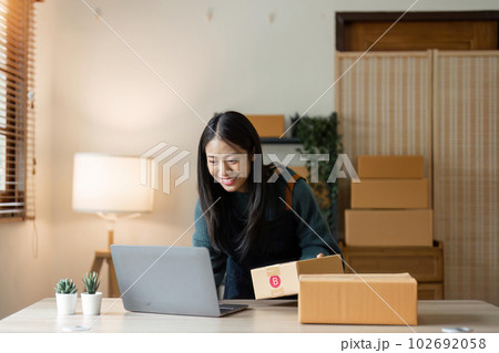 Asian businesswoman using laptop to start online delivery freelancer or salesman checking production order with laptop. SME entrepreneur working with parcel box at home Asian businesswoman using laptop to start online delivery freelancer or salesman checking production order with laptop. SME entrepreneur working with parcel box at home 102692058